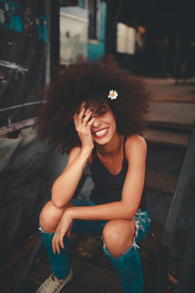 Current image: Cheerful woman wearing ripped jeans and curly hair with flower, enjoying a sunny day outdoors.
